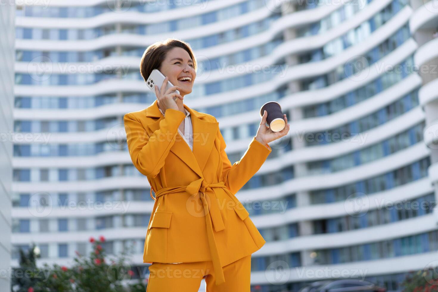 Smiling  beautiful  woman with short haircut  in yellow jacket and pants using mobyle phone , holding cup of coffe . Standing over modern city. photo