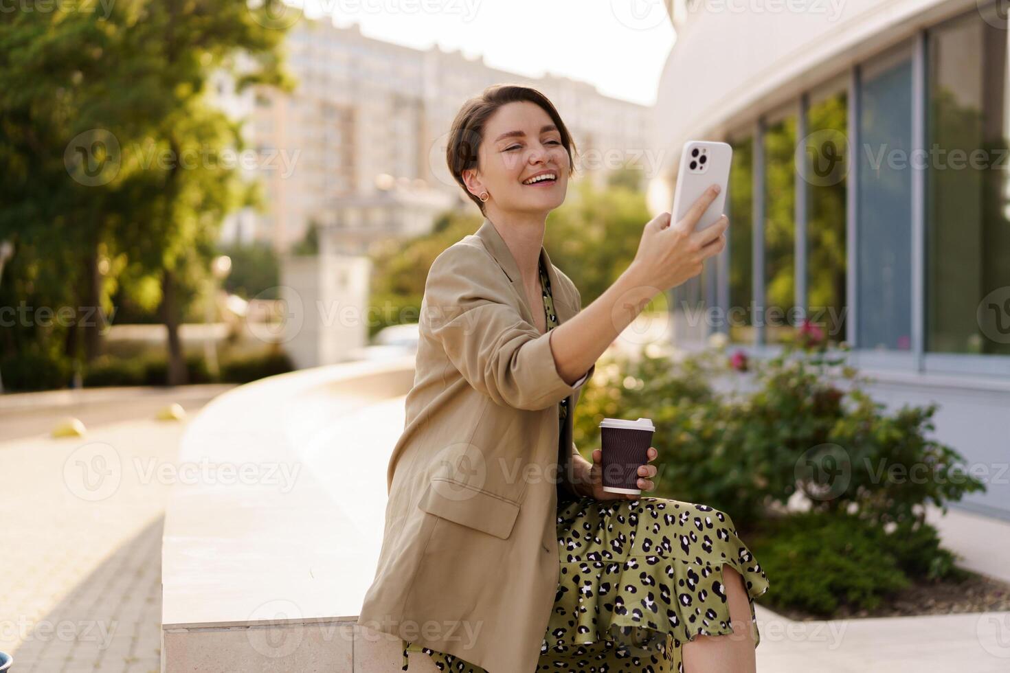Outdoor photo of stylish beautiful woman with short haircut  enjoing autu,n day  holding cup of coffee, using mobyle phone. Weaering casual  jacker and elegant dress.