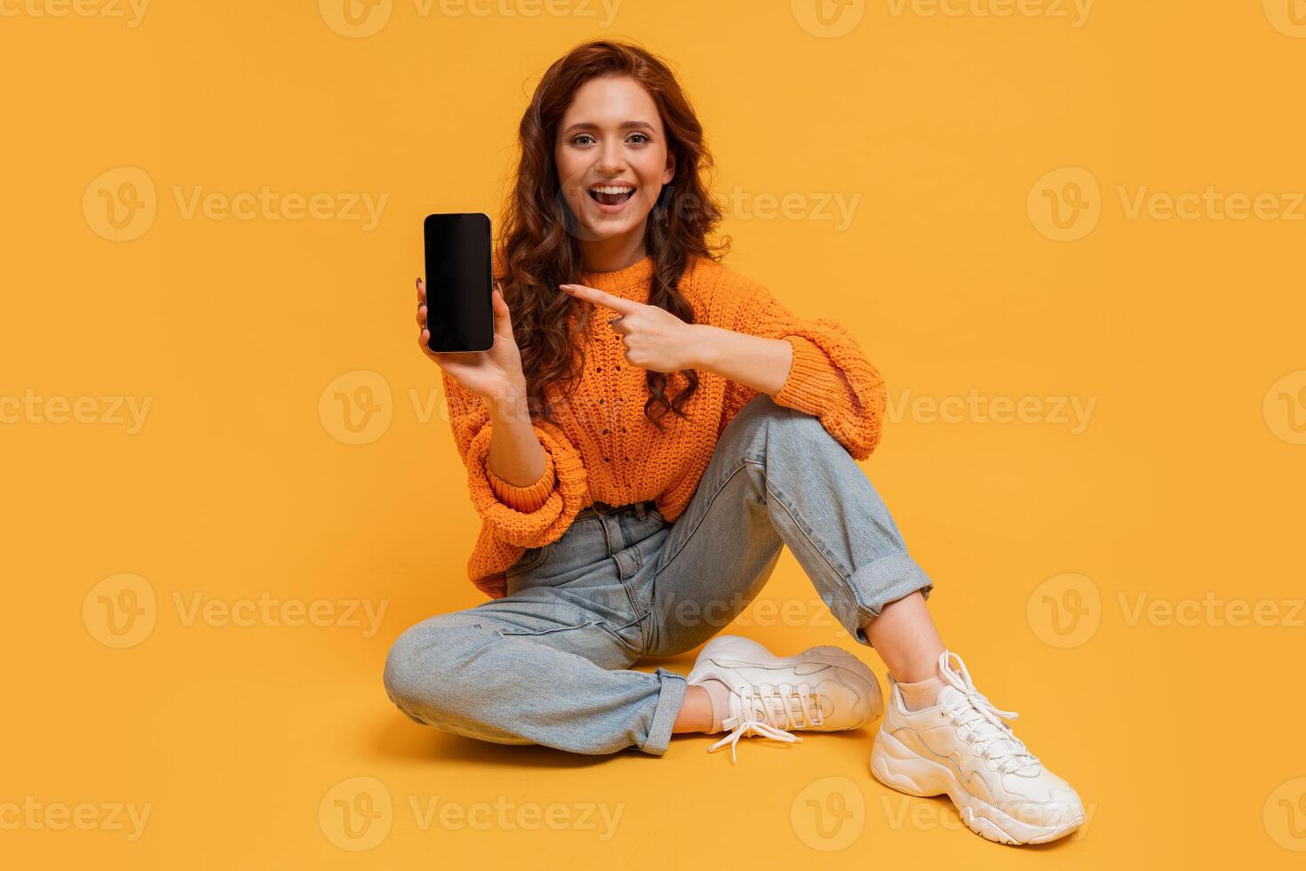 Happy millennial woman showing smartphone screen and pointing on it.  Wearing trendy autumn autfit. Studio photo. photo
