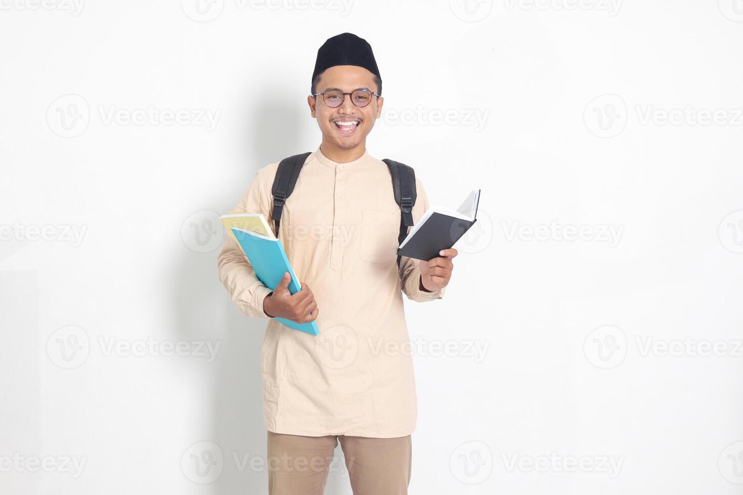 Portrait of excited student Asian muslim man in koko shirt with skullcap carrying school books, while reading his notebook. Isolated image on white background photo