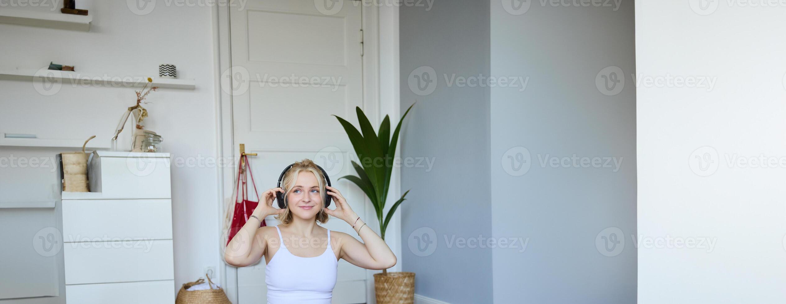 Vertical shot of smiling young woman using tutorials for workout at home, sitting with laptop in wireless headphones on yoga mat, follows fitness instructor on social media, repeats exercises photo
