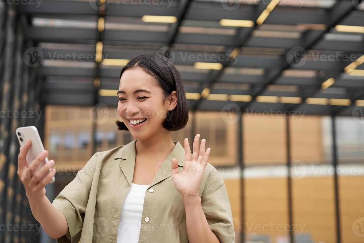 Happy asian girl waves at her smartphone camera, says hello to friend on chat, calling someone, standing on street photo