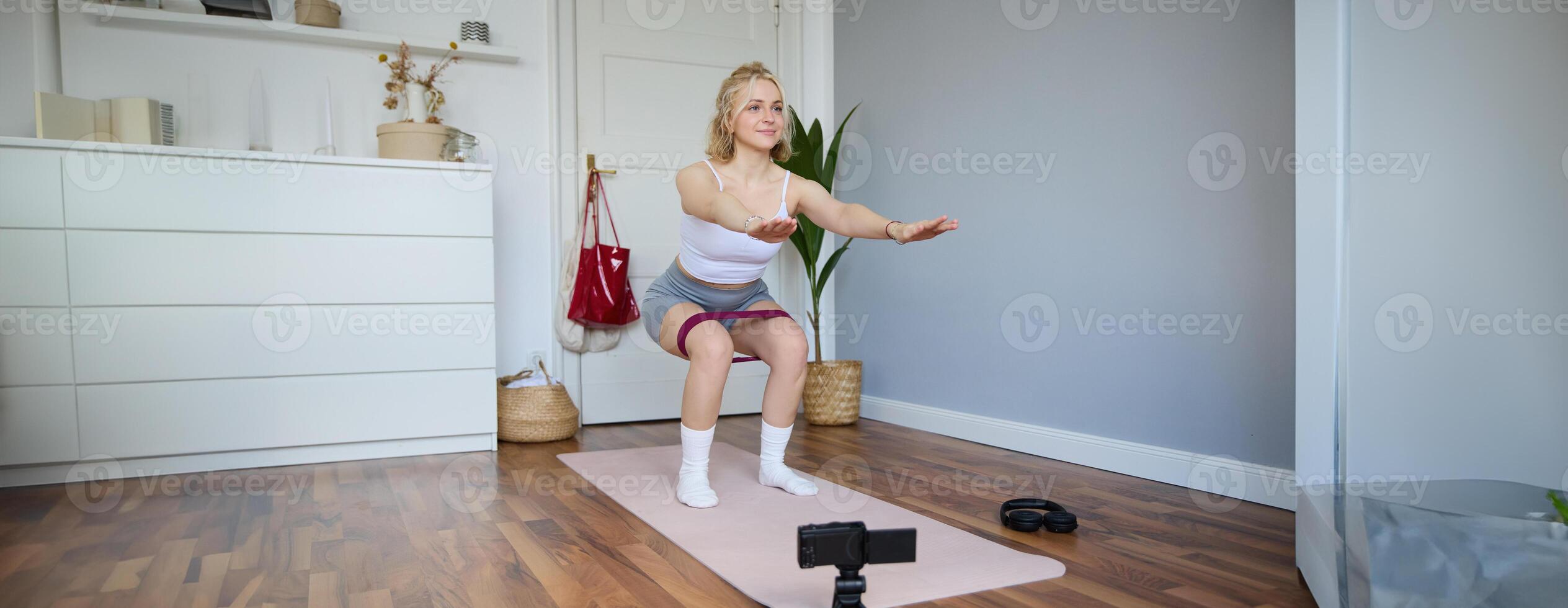 Portrait of young woman, fitness instructor making a for sport vlog, doing squats on camera, using resistance band, working out at home photo