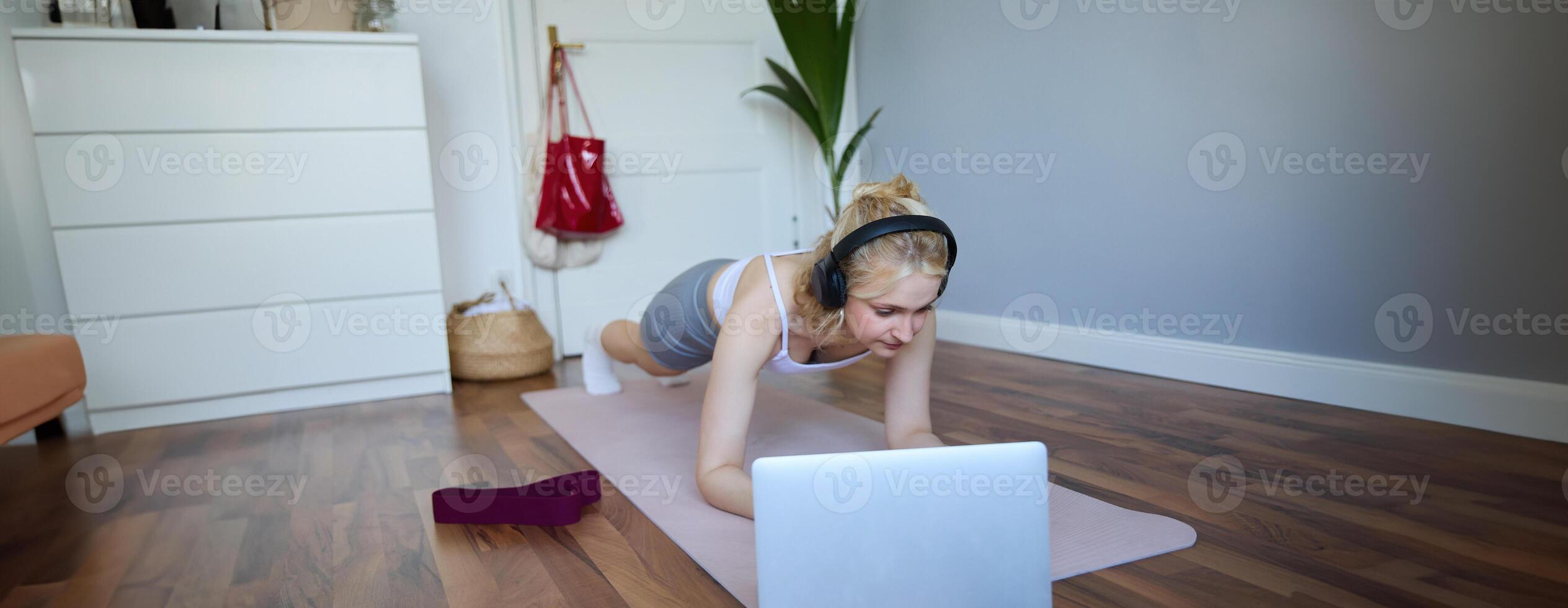 Portrait of young athletic woman in wireless headphones, standing in plank, following online workout , using laptop to join training session on the internet photo