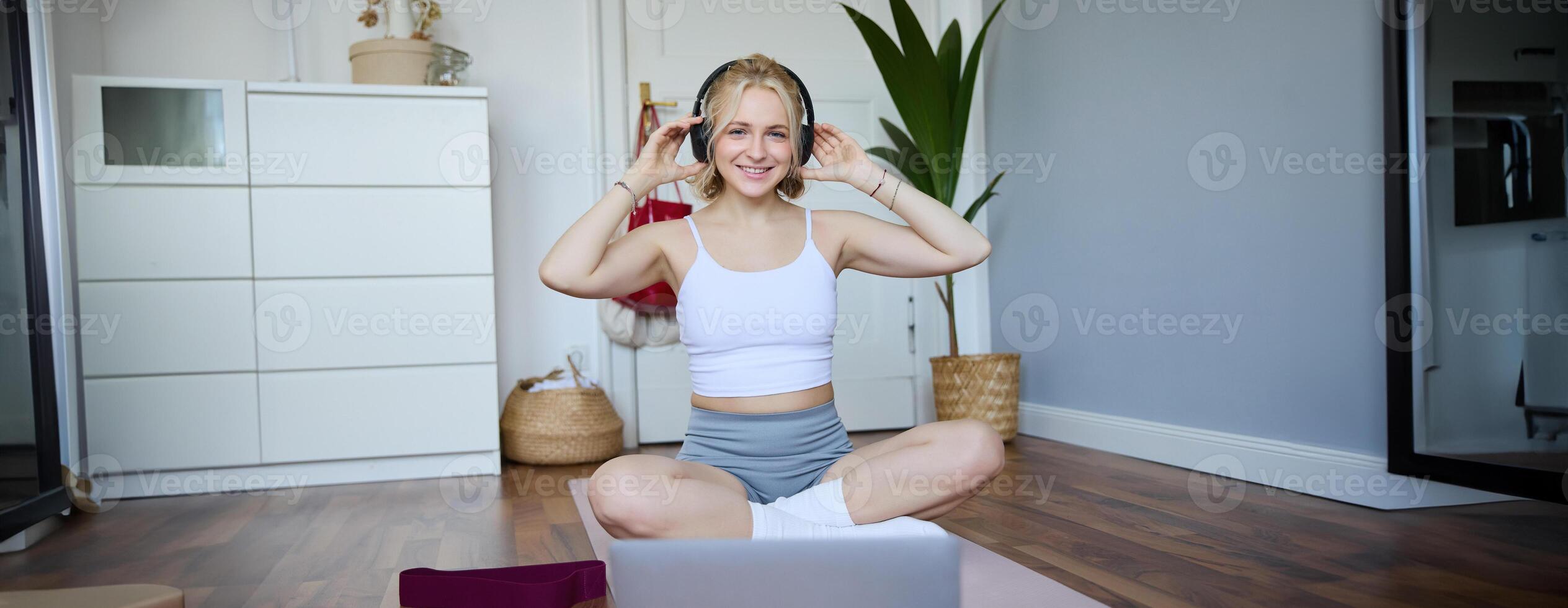 Portrait of cute young fitness woman, connects to online training session via laptop, wearing wireless headphones during workout, sits at home on rubber yoga mat photo