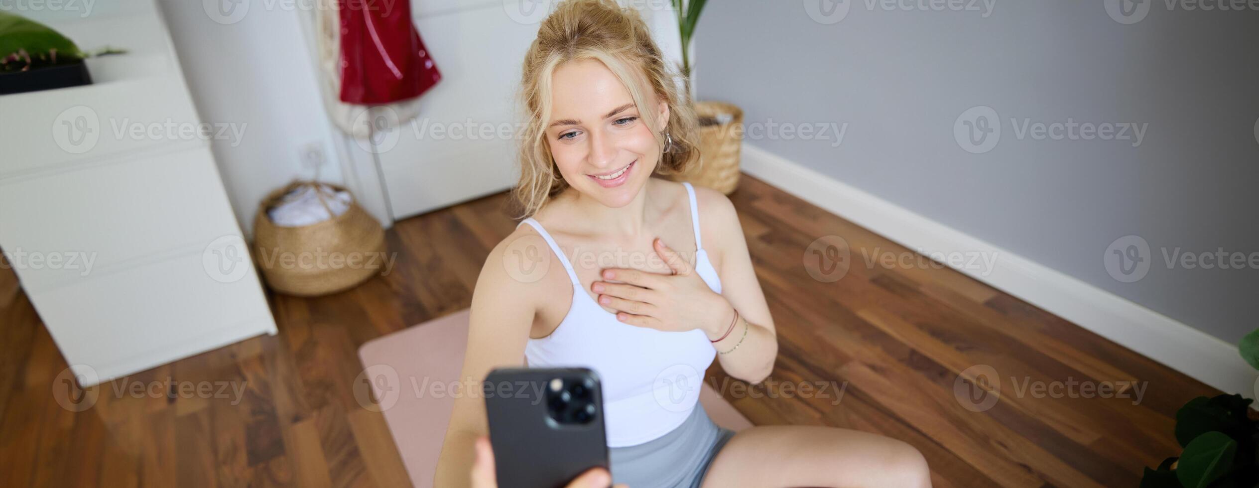Portrait of beautiful young fitness trainer, showing home workout, taking selfie on mobile phone while sitting on yoga rubber mat in bedroom, wearing activewear photo