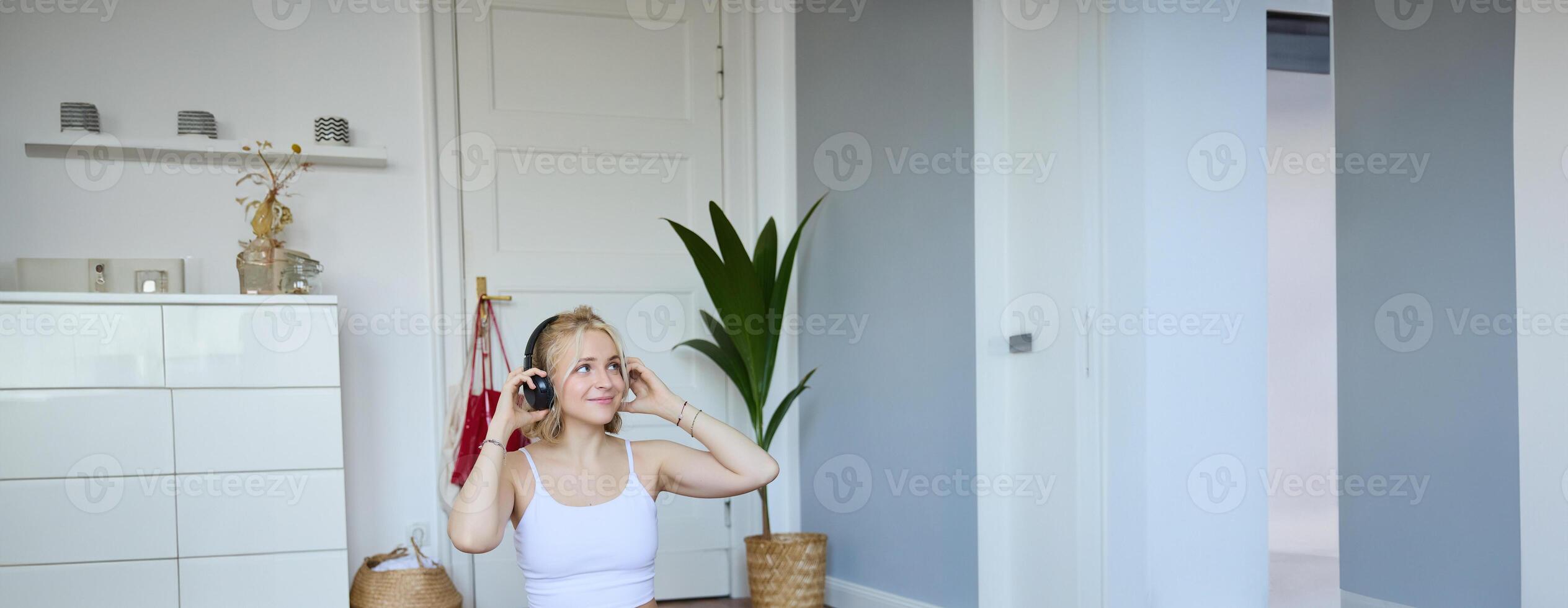 Vertical shot of smiling young woman using tutorials for workout at home, sitting with laptop in wireless headphones on yoga mat, follows fitness instructor on social media, repeats exercises photo