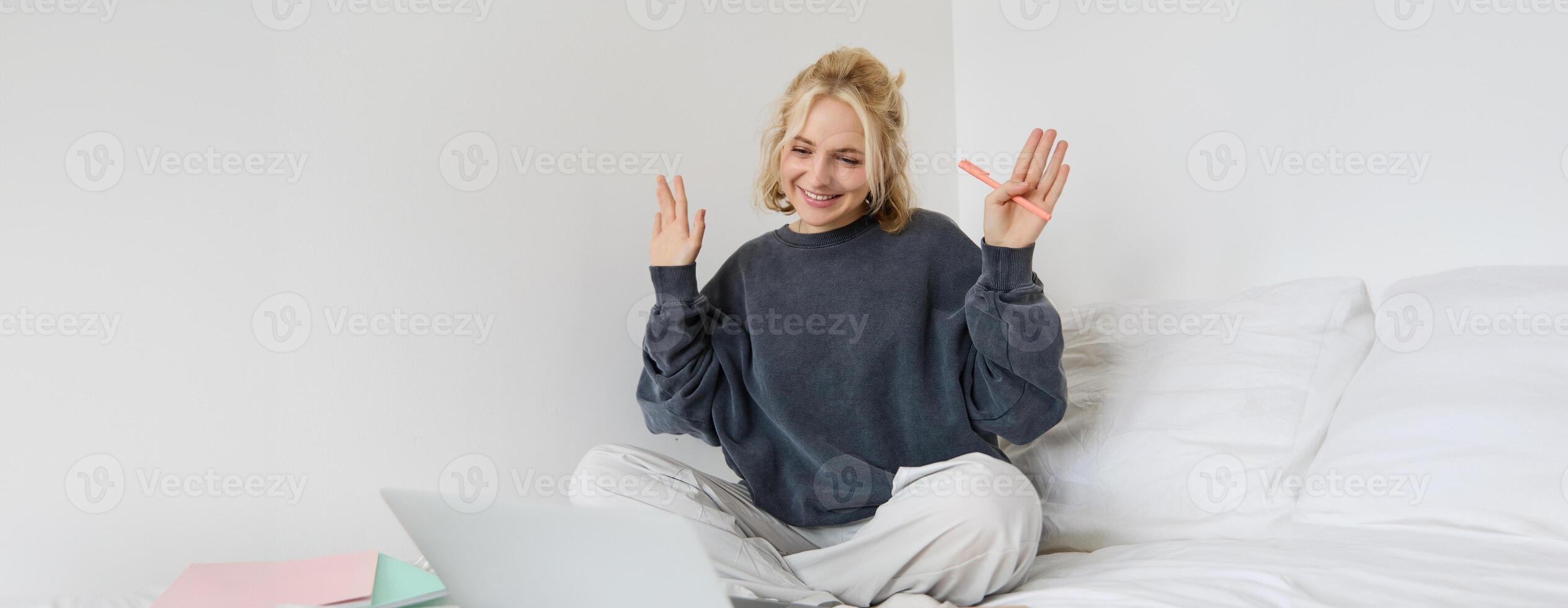 Portrait of young happy woman, connects to chat, using laptop, waving hand at camera, saying hello to someone online, sitting on bed, studying, e-learning or working from home photo
