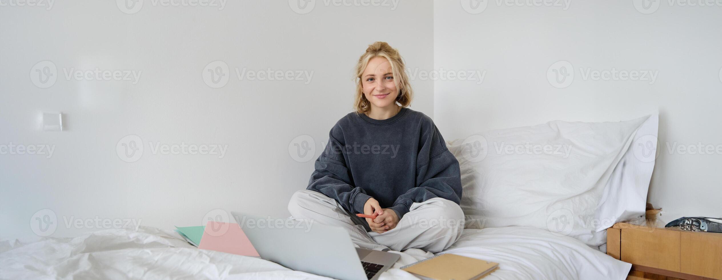 Portrait of happy blond woman, freelancer working from home, sitting on bed with laptop and notebooks. Student doing homework in bedroom, connects to online class via chat photo
