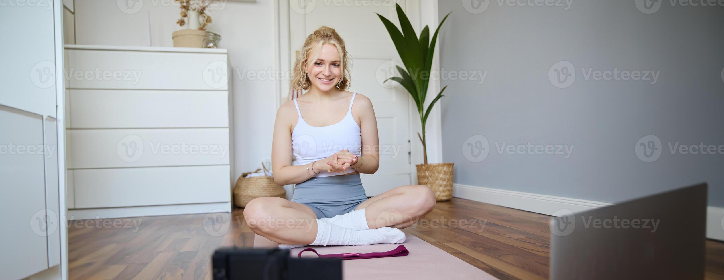 Portrait of young charismatic fitness trainer, girl blogger records on digital camera, talks about health and workout, doing exercises on rubber mat in a room at home photo