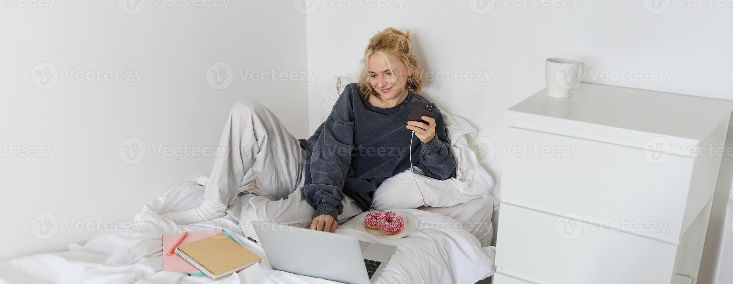 Lifestyle and weekend concept. Young smiling woman, relaxing at home, lying in bed with cup of tea and doughnut, using laptop and smartphone in bedroom photo