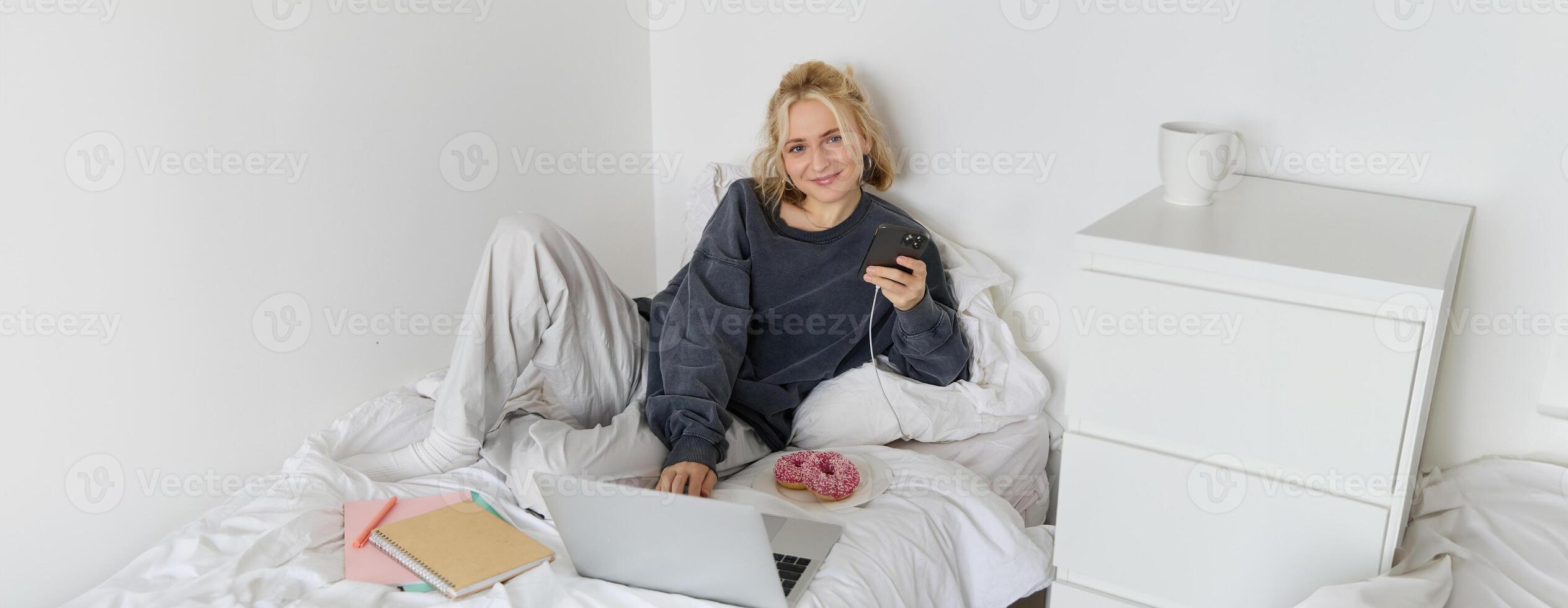 Portrait of young woman, student studying in her bed, relaxing while preparing homework, eating doughnut, using laptop in bedroom and drinking tea photo