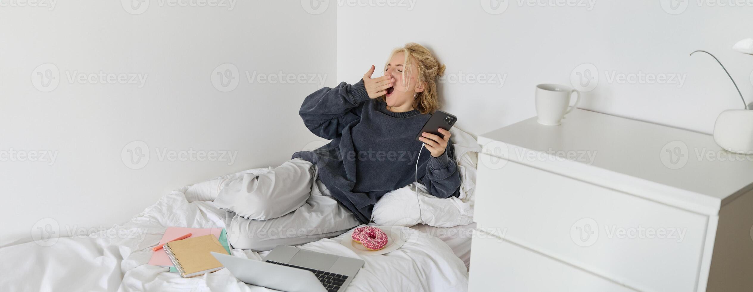Portrait of young woman spending lazy weekend at home, lying in bed with smartphone and laptop, eats doughnut and yawns, feels sleepy or bored photo