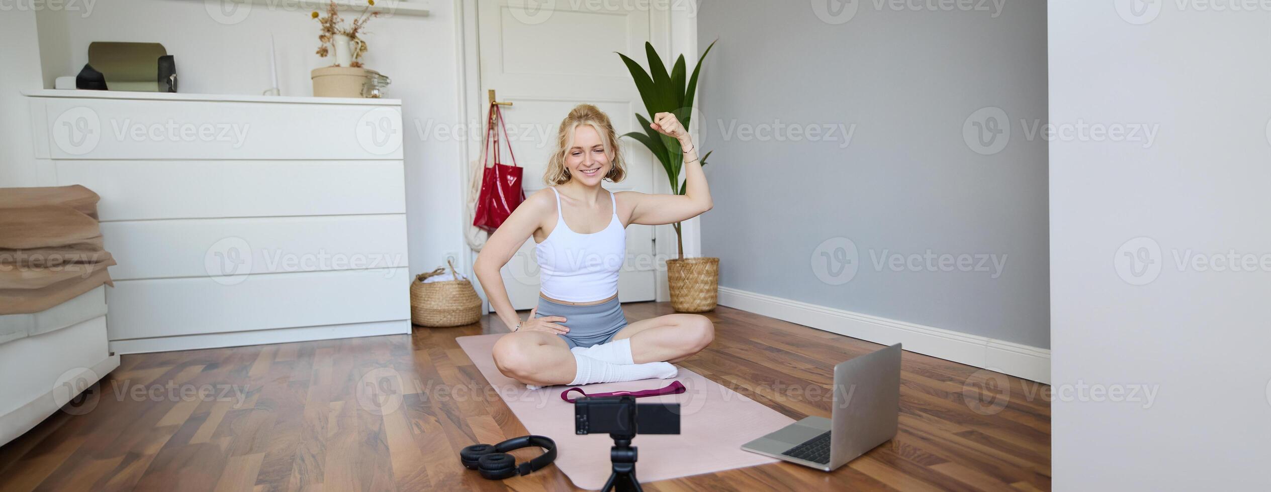 Portrait of young female athlete, fitness trainer recording vlog, training session on digital camera, sitting in a room on rubber yoga mat, showing exercises photo