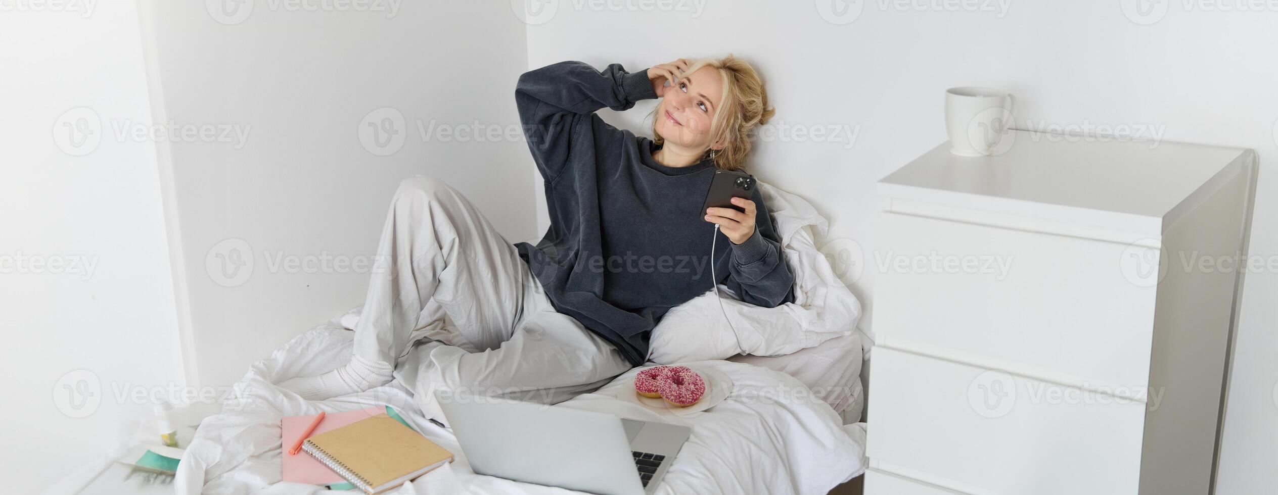 Lifestyle and weekend concept. Young smiling woman, relaxing at home, lying in bed with cup of tea and doughnut, using laptop and smartphone in bedroom photo