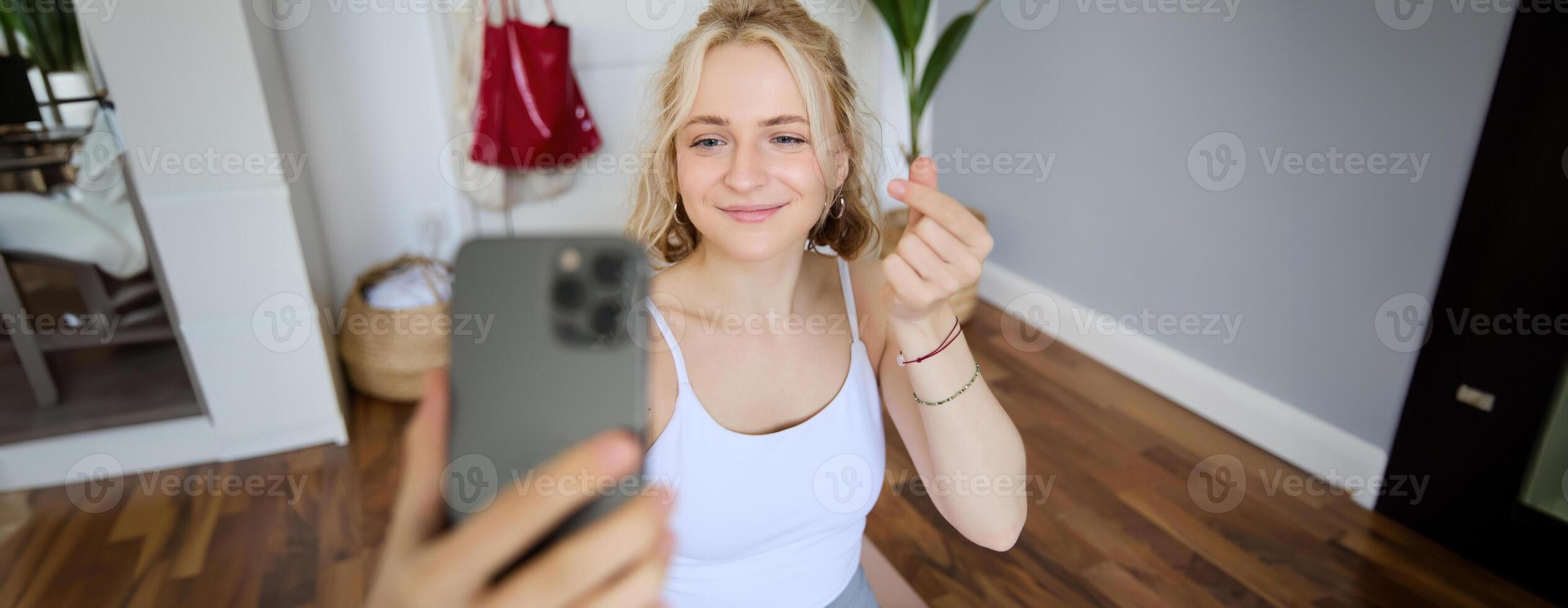 Portrait of young beautiful blond woman, fitness instructor doing exercises at home on yoga mat, taking selfies on smartphone, recording of herself for social media account about workout photo