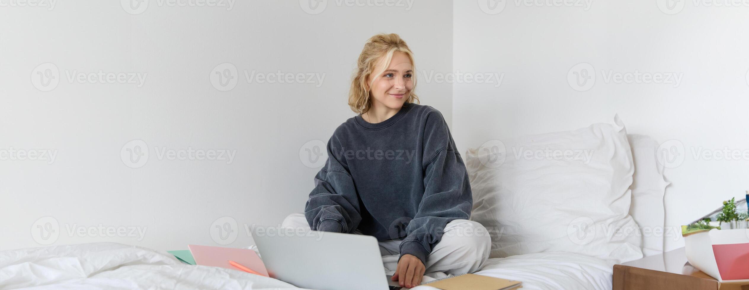 Portrait of happy blond woman, freelancer working from home, sitting on bed with laptop and notebooks. Student doing homework in bedroom, connects to online class via chat photo