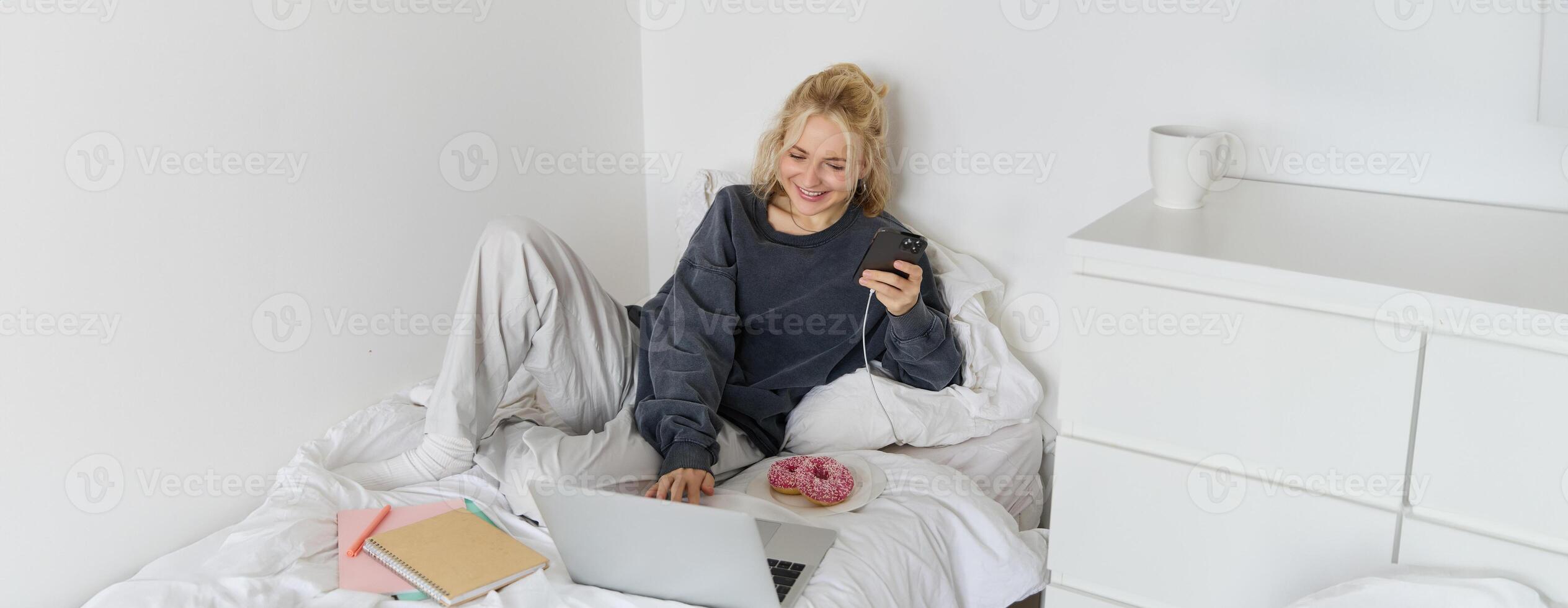 Portrait of smiling candid woman, lying in bed with doughnut, using smartphone and laptop, resting at home in bedroom, watching tv show or chatting online photo