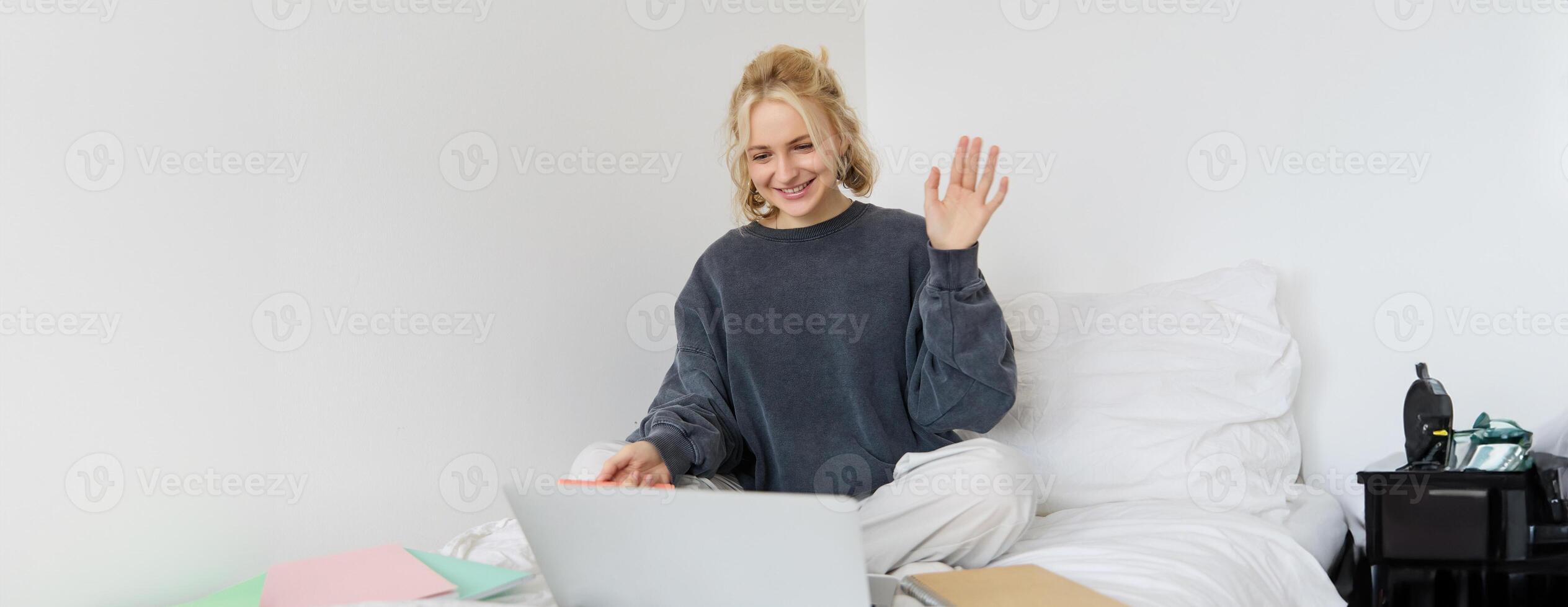 Portrait of young happy woman, connects to chat, using laptop, waving hand at camera, saying hello to someone online, sitting on bed, studying, e-learning or working from home photo