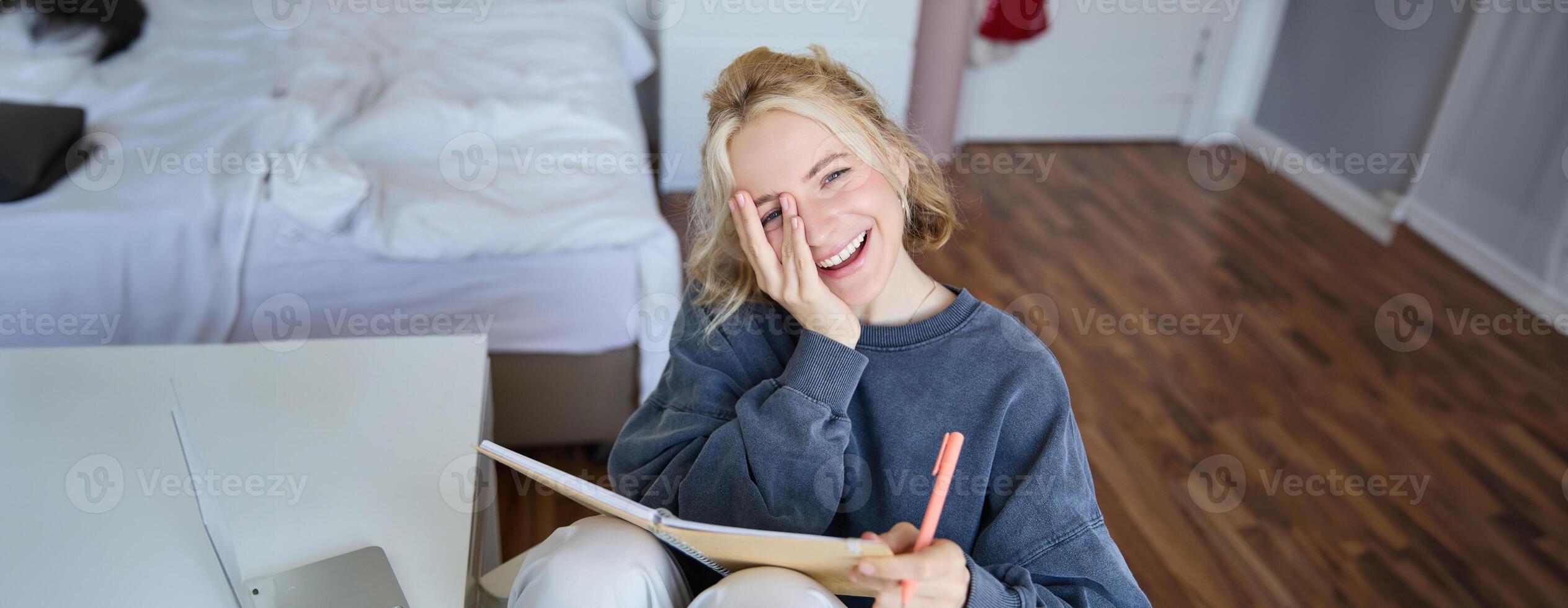 Portrait of happy, young blond woman, sitting in bedroom with notebook and pen, laughing and smiling, writing in journal, doing homework and smiling photo