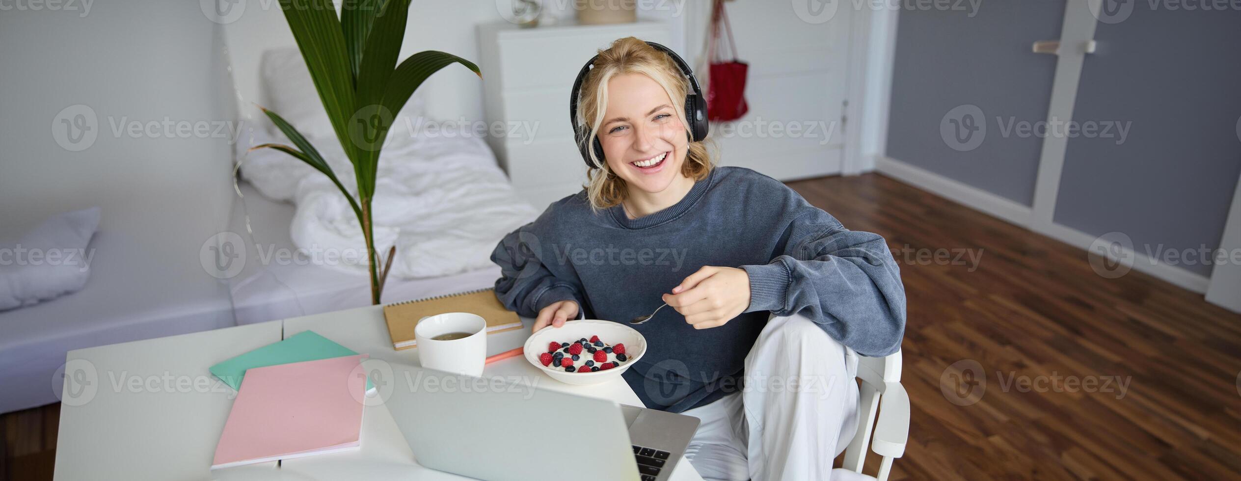 Image of happy woman sitting in a room, watching interesting tv show or movie on laptop, using screaming service, wearing headphones, eating dessert and drinking tea photo