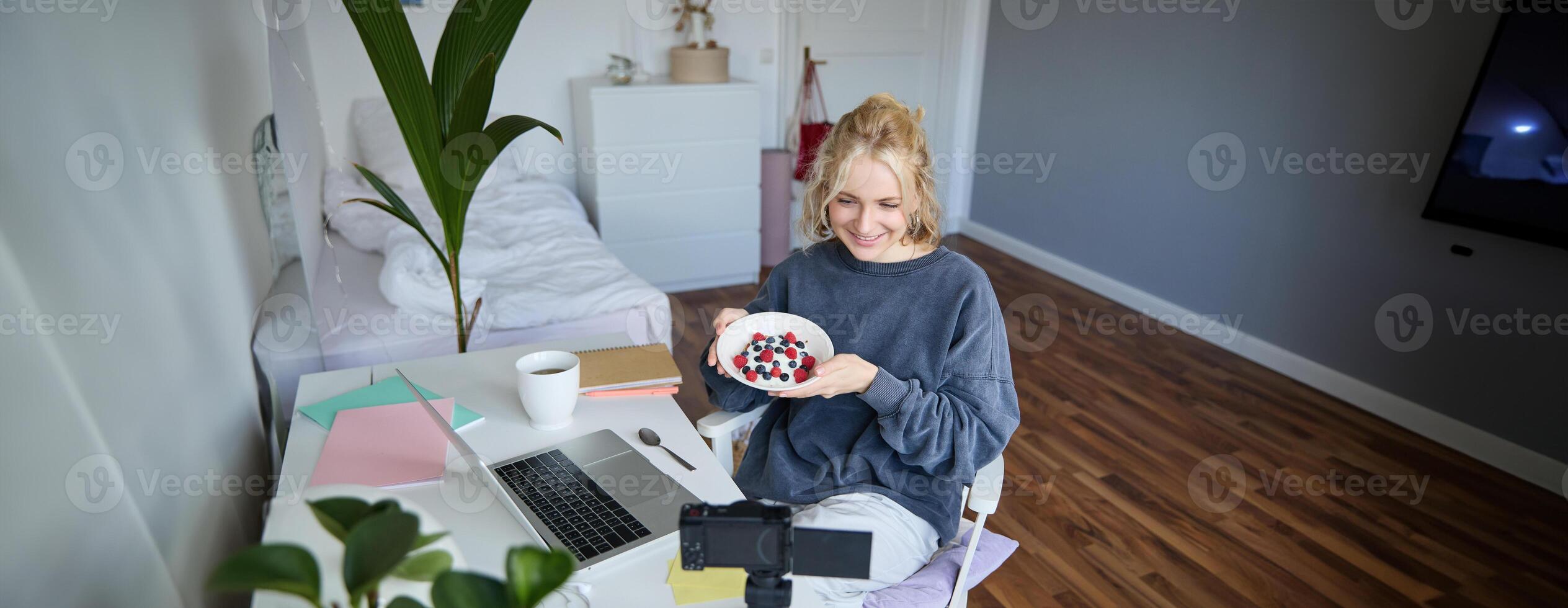 Portrait of cute young girl vlogger, showing her breakfast on camera, recording vlog in her bedroom, holding dessert, eating photo