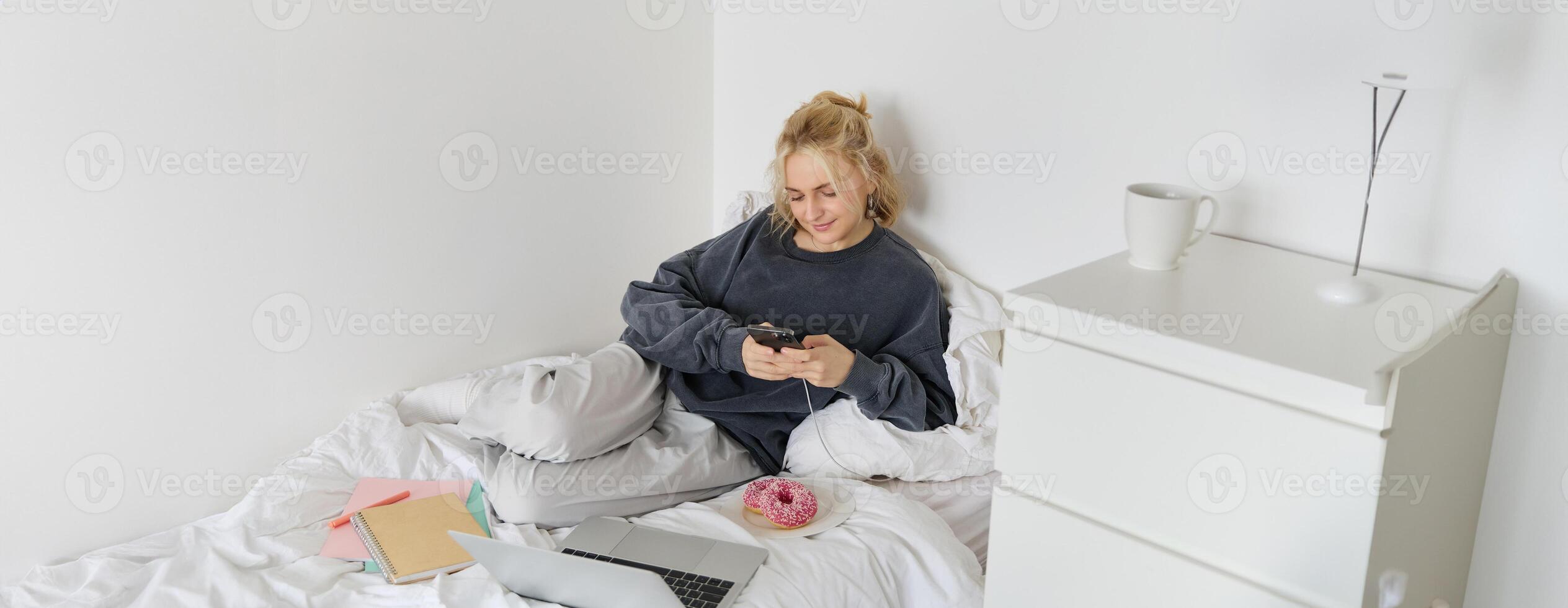 Lifestyle and people concept. Young beautiful woman, staying at home, lying in bed with laptop and smartphone, eating doughnut, enjoying free time, spending weekend at home, watching movie online photo