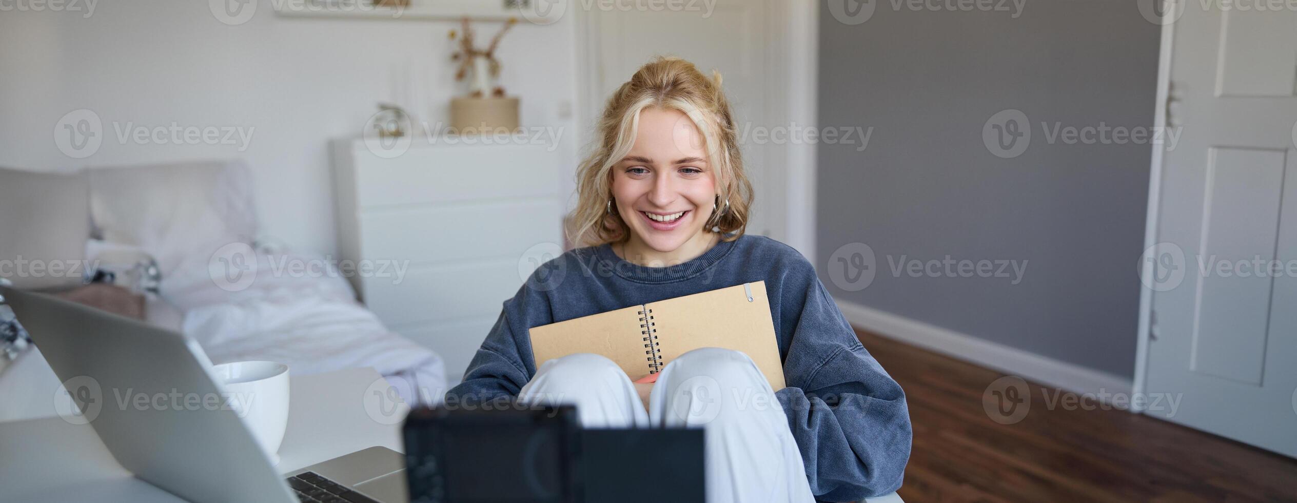 Portrait of blond smiling woman, records on digital camera how she writes in notebook, talks to followers, doing lifestyle blog content in her room photo