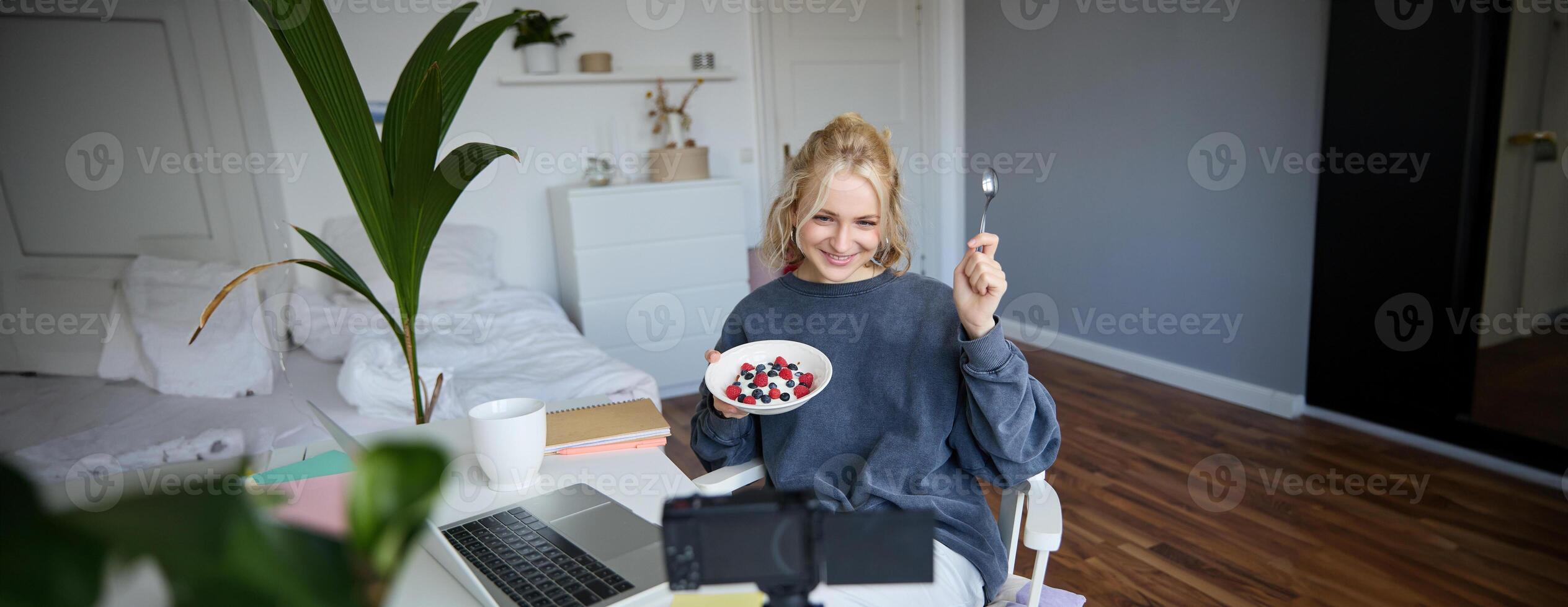 Portrait of cute young girl vlogger, showing her breakfast on camera, recording vlog in her bedroom, holding dessert, eating photo