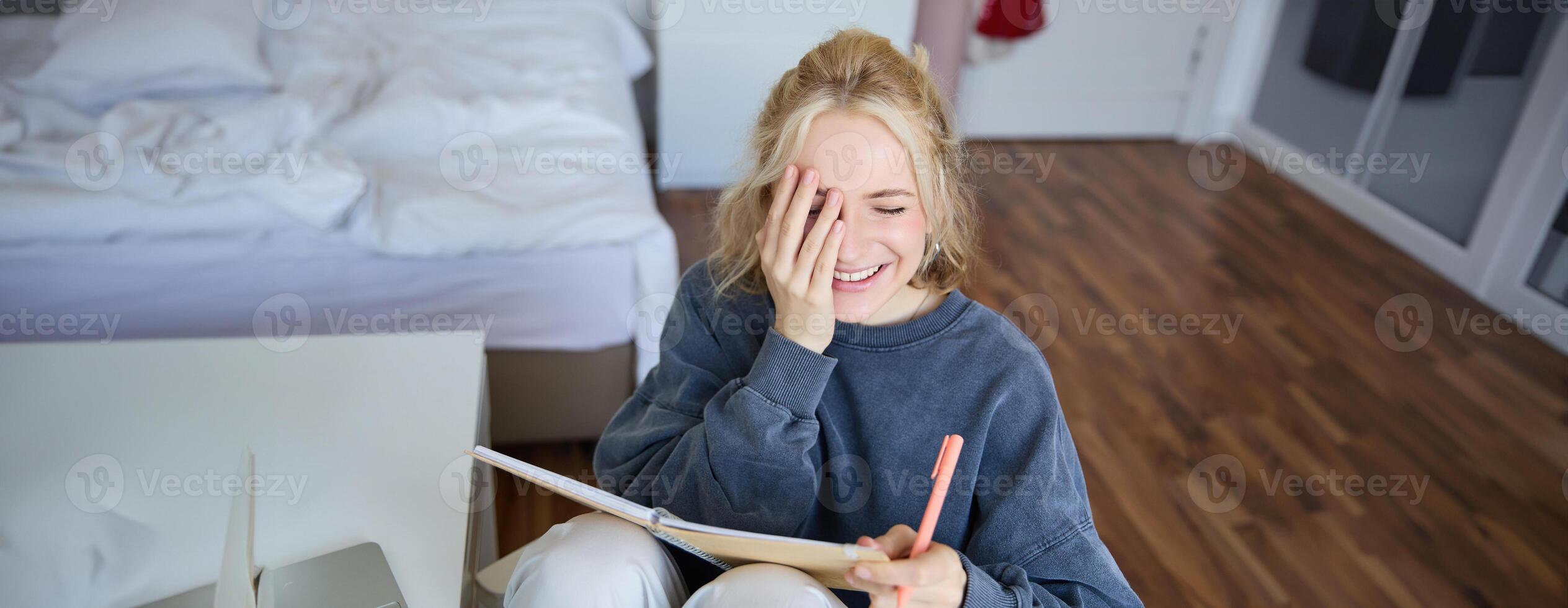 Portrait of charismatic blond girl, smiling woman in bedroom, holding notebook and pen, writing in journal or diary, creates to do list in planner photo