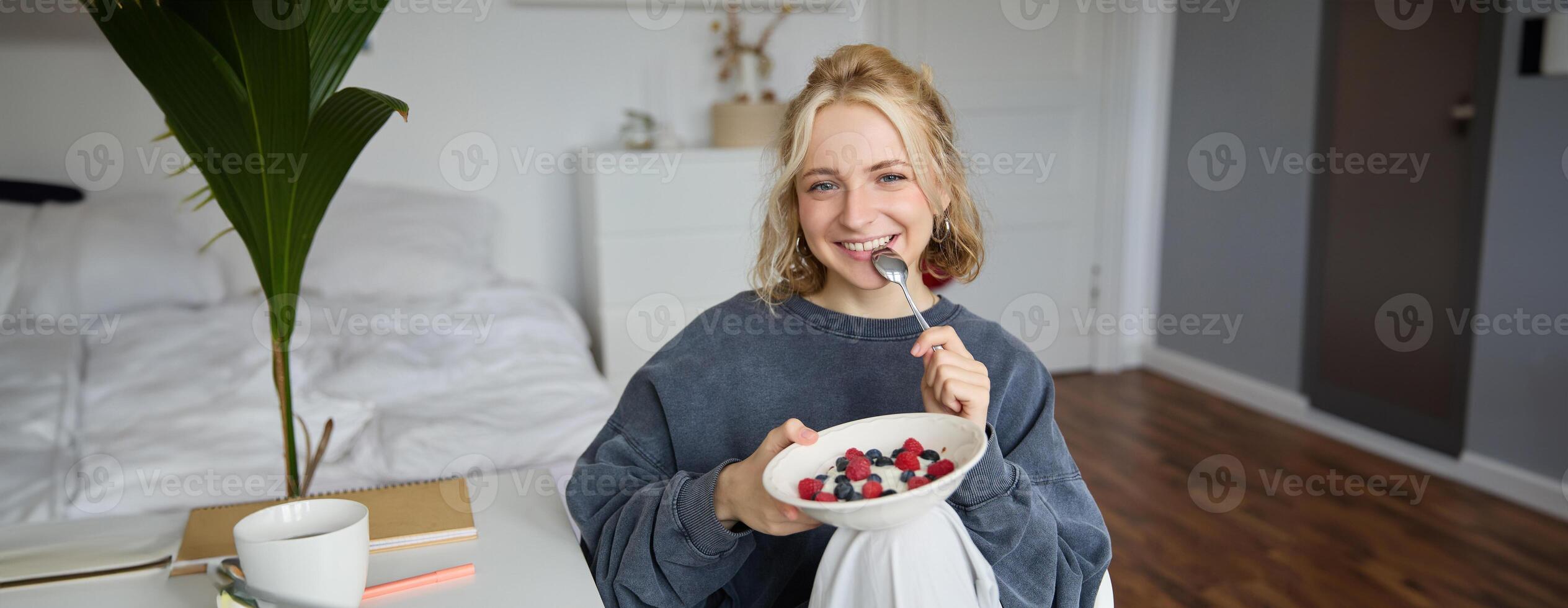 Portrait of smiling blond woman, eating breakfast, holding bowl and spoon, sitting in bedroom, looking happy at camera photo