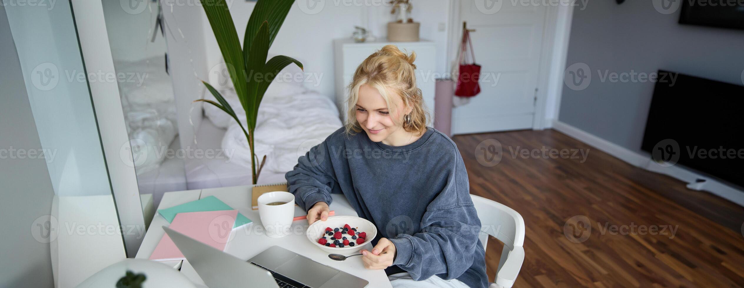 Portrait of smiling blond young woman, eating in front of laptop, watching s online while having breakfast, enjoying dessert, sitting in bedroom photo