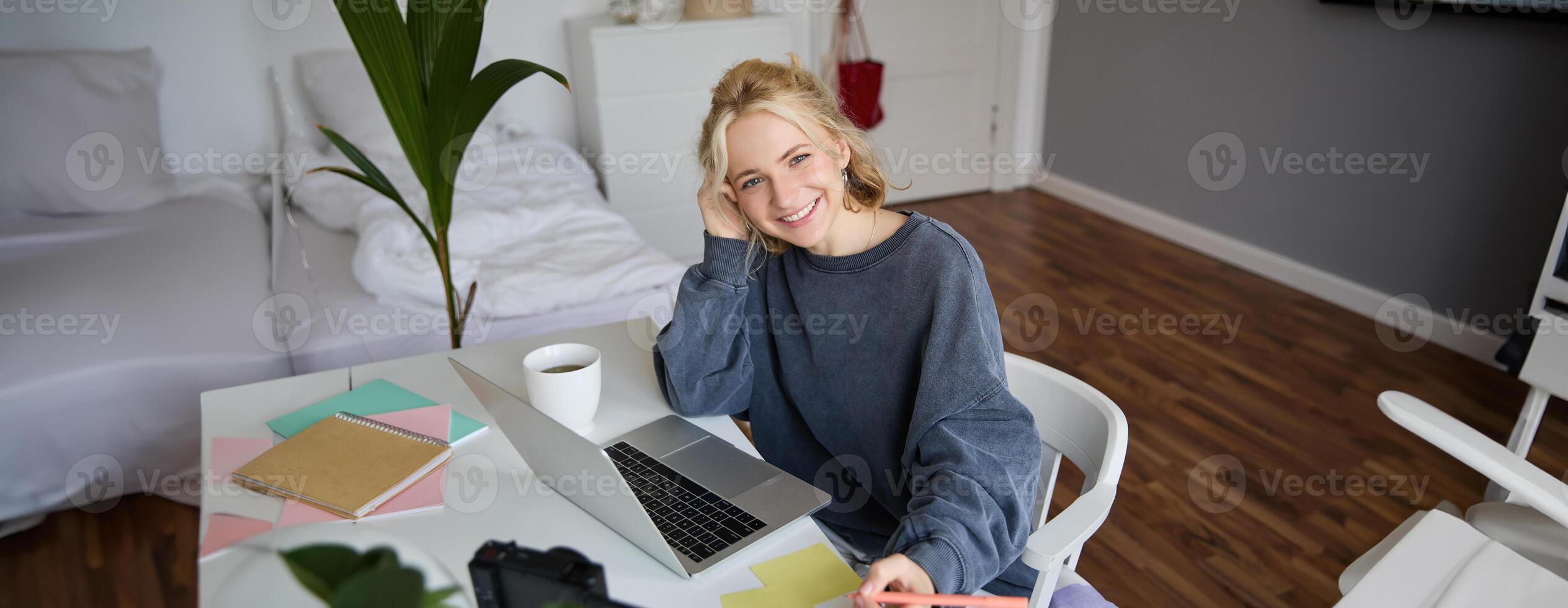 Portrait of smiling, beautiful blond woman, writing down notes, doing homework, studying from home, doing distance learning, online course, working remotely in her room photo