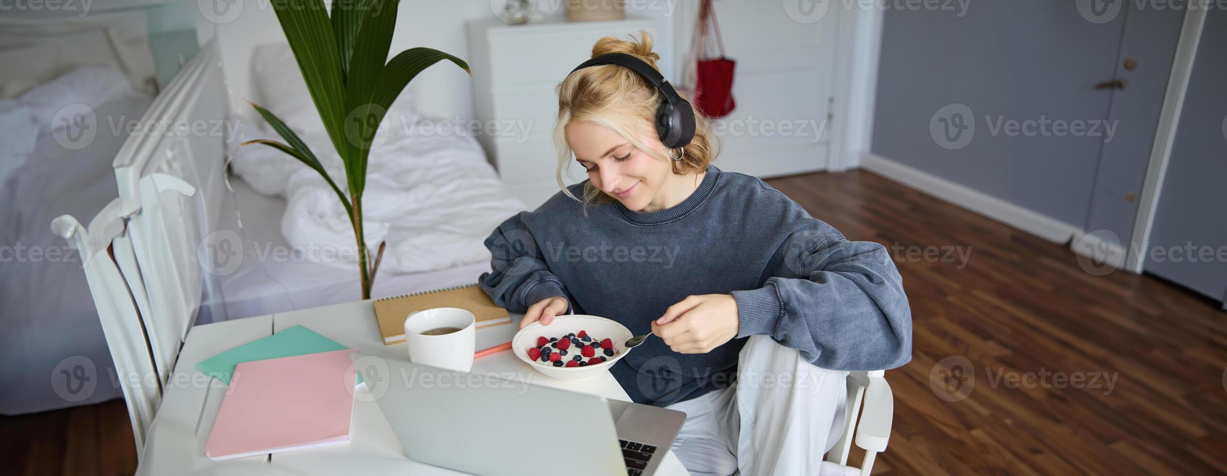 Portrait of smiling young woman, watching tv show in headphones, eating breakfast and looking at laptop screen photo