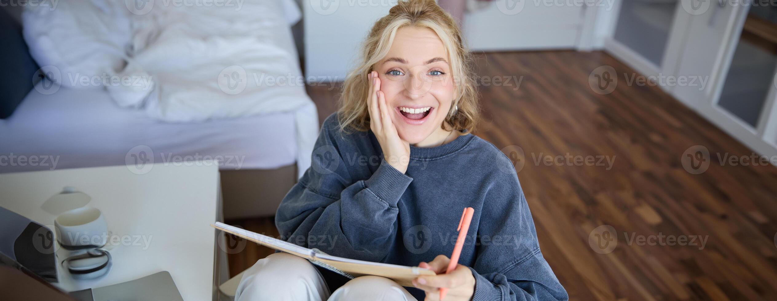 Portrait of happy, young blond woman, sitting in bedroom with notebook and pen, laughing and smiling, writing in journal, doing homework and smiling photo