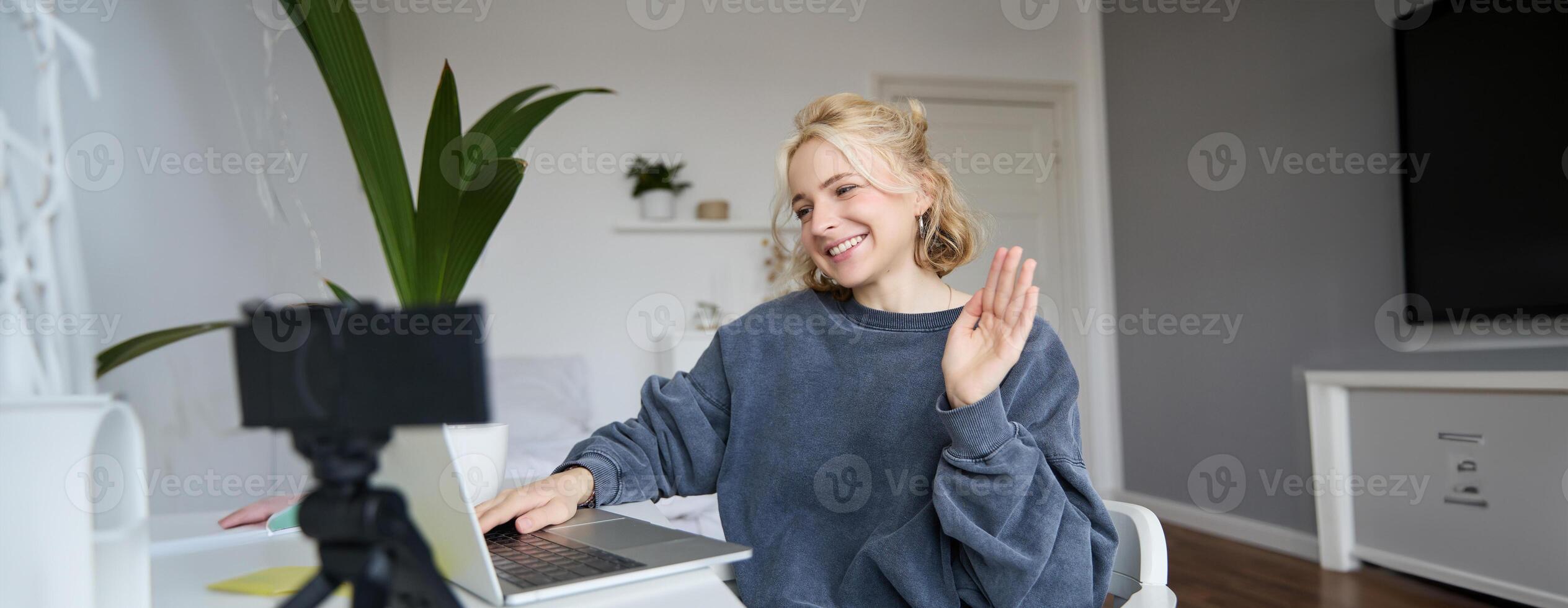 Image of smiling beautiful young woman, chats online on laptop, waves hand, says hello, records on digital camera, vlogging, creates content for her blog photo