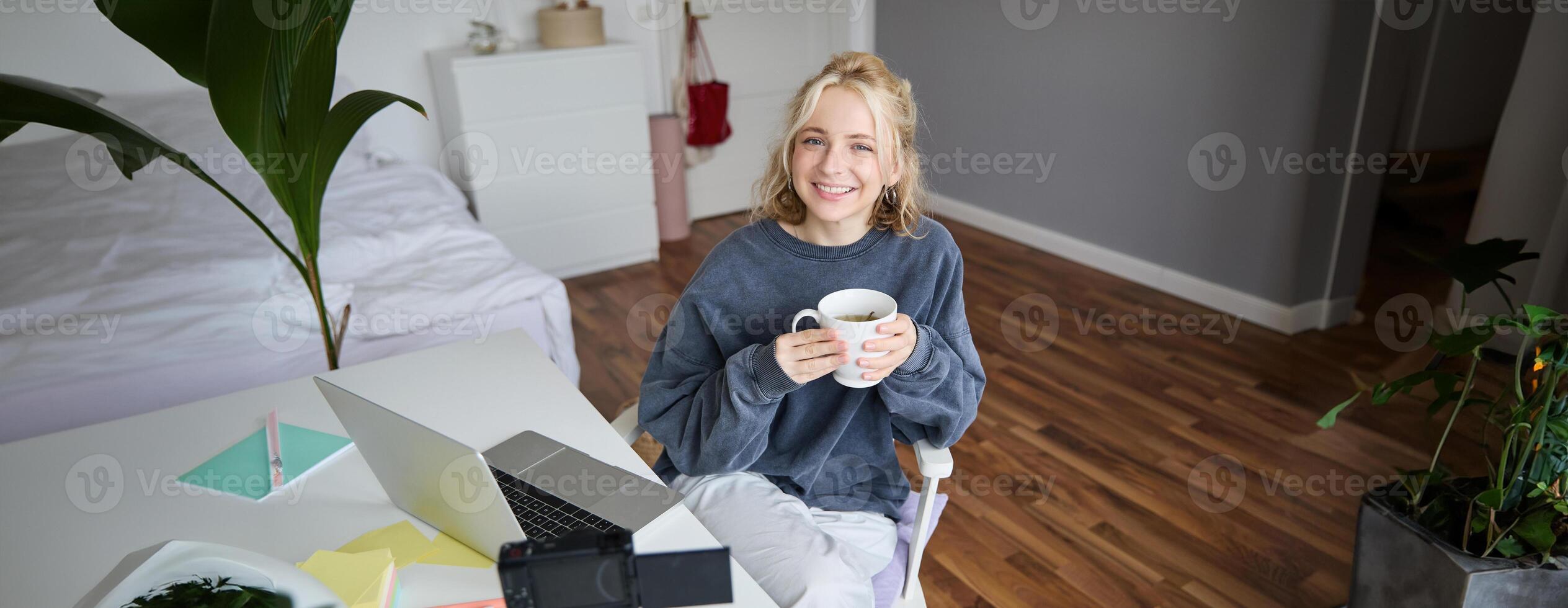 Image of young woman, social media influencer, editing her on laptop, sits in a room with computer and digital camera, drinking coffee, smiling at camera photo