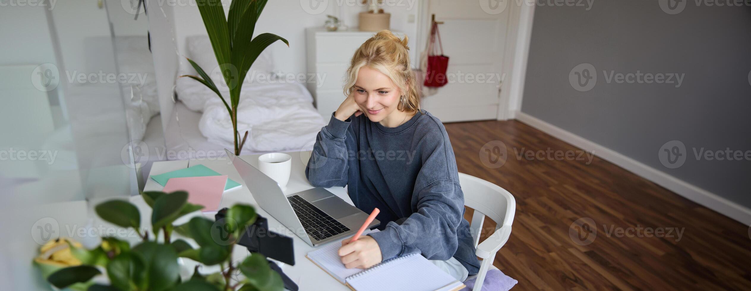 Portrait of smiling, beautiful blond woman, writing down notes, doing homework, studying from home, doing distance learning, online course, working remotely in her room photo