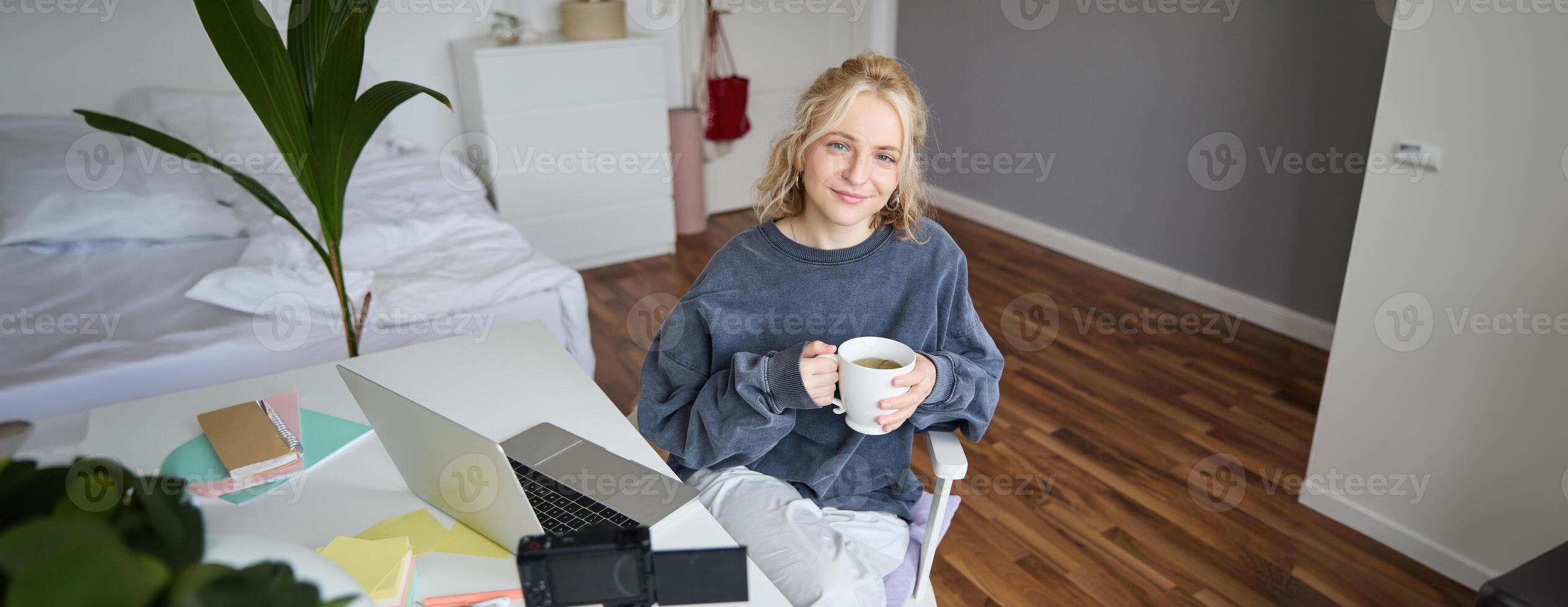 Image of young woman, social media influencer, editing her on laptop, sits in a room with computer and digital camera, drinking coffee, smiling at camera photo
