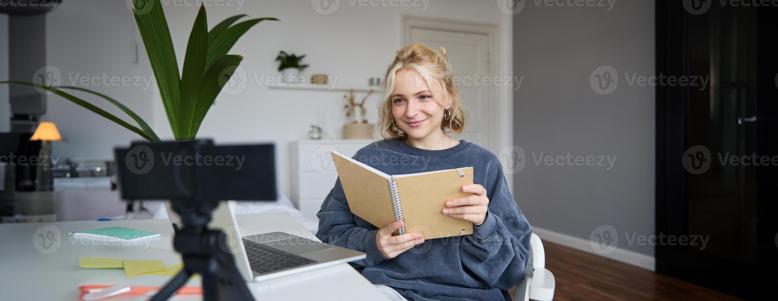 Young smiling blond woman, sits near laptop, uses digital camera to record blog, creates lifestyle content for social media, records tutorial, shows notebook, reads her notes photo