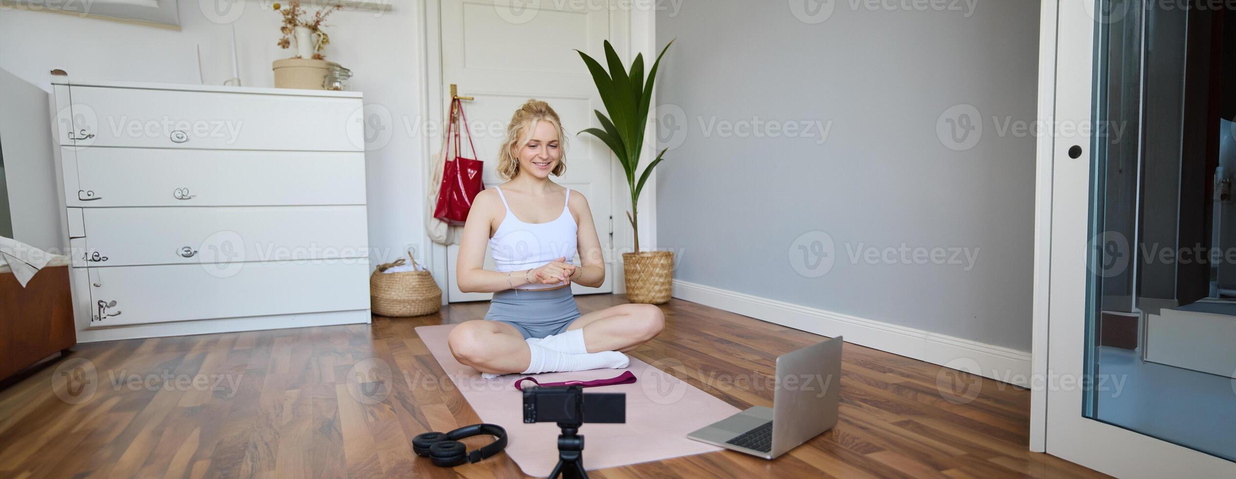 Portrait of young fitness blogger, woman showing exercises to her followers, recording on digital camera, doing workout training session, home yoga, sitting in front of laptop on rubber mat photo