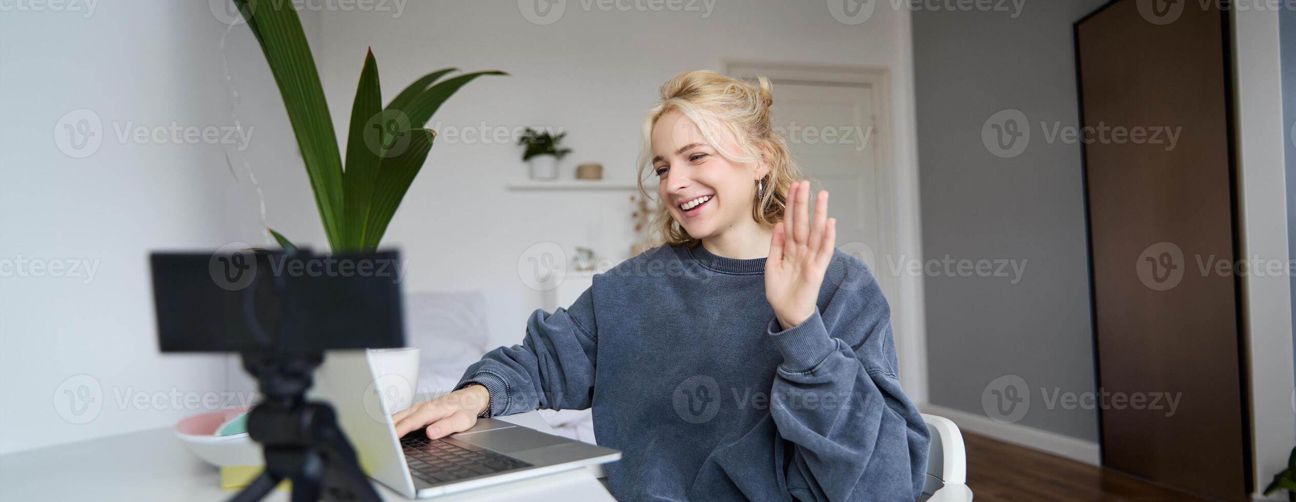 Portrait of young social media content creator, woman sits in room with laptop and digital camera, records lifestyle vlog, says hello, chats to someone online via computer photo