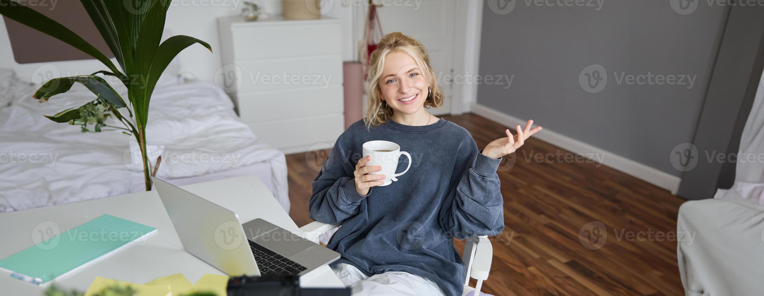 Image of young woman, social media influencer, editing her on laptop, sits in a room with computer and digital camera, drinking coffee, smiling at camera photo