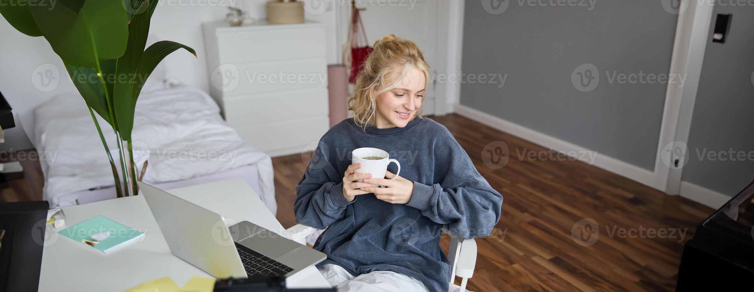 Portrait of young smiling woman, vlogger recording lifestyle in her room, sits in front of laptop and digital camera, drinks tea, creates content for social media account photo