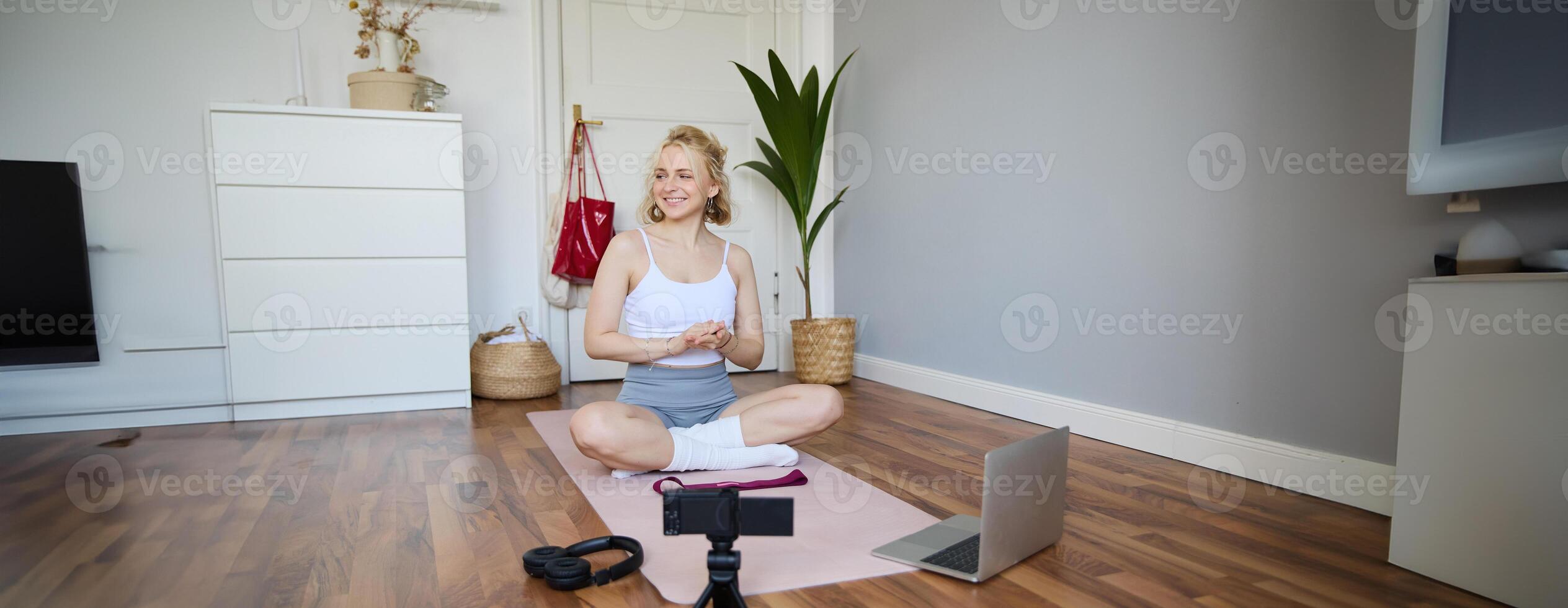 Portrait of young fitness blogger, woman showing exercises to her followers, recording on digital camera, doing workout training session, home yoga, sitting in front of laptop on rubber mat photo