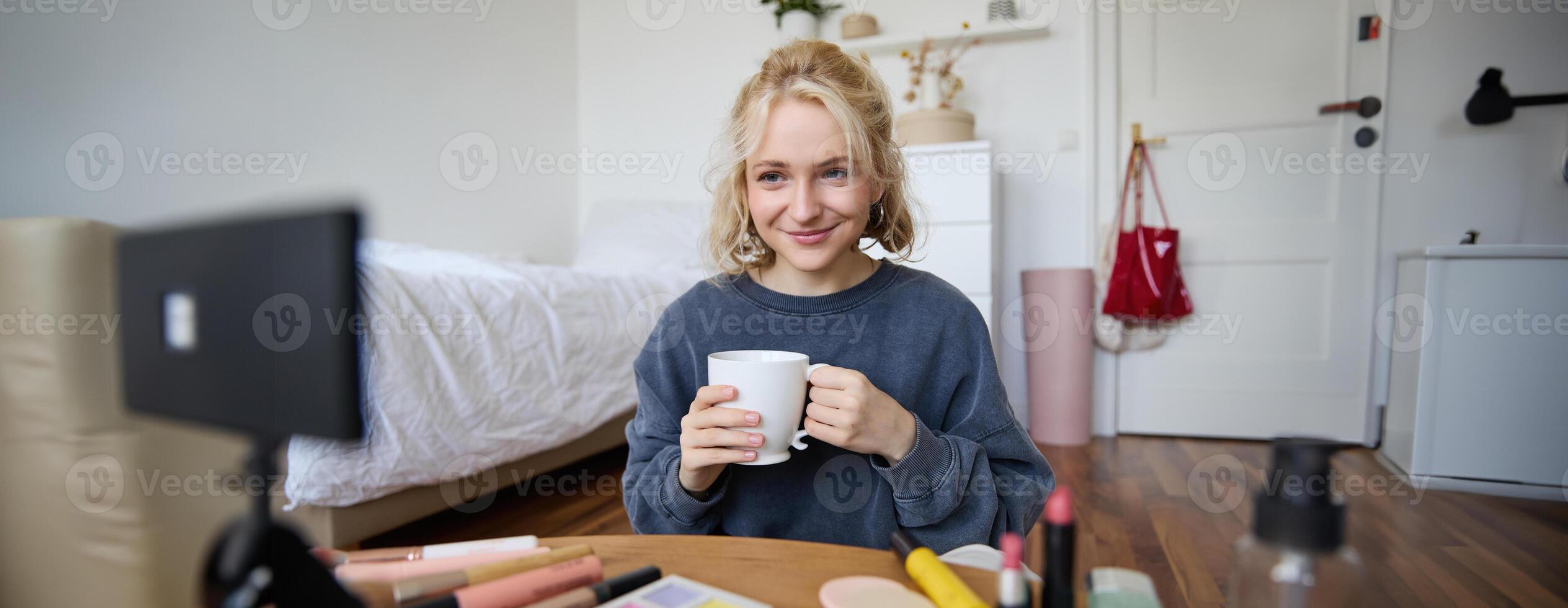 Image of young woman, makeup vlogger, sitting in bedroom with digital camera, drinking tea and talking, creating lifestyle , social media content photo