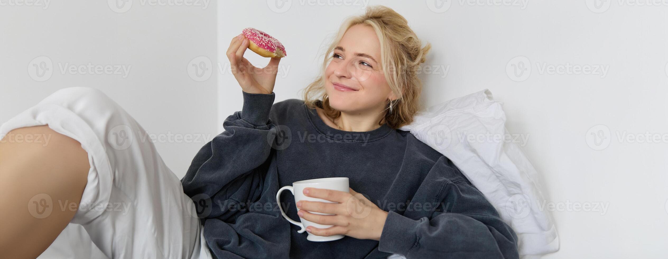 Close up portrait of beautiful smiling, blond woman, lying in bed, drinks tea and eats doughnut on a lazy weekend or day-off photo