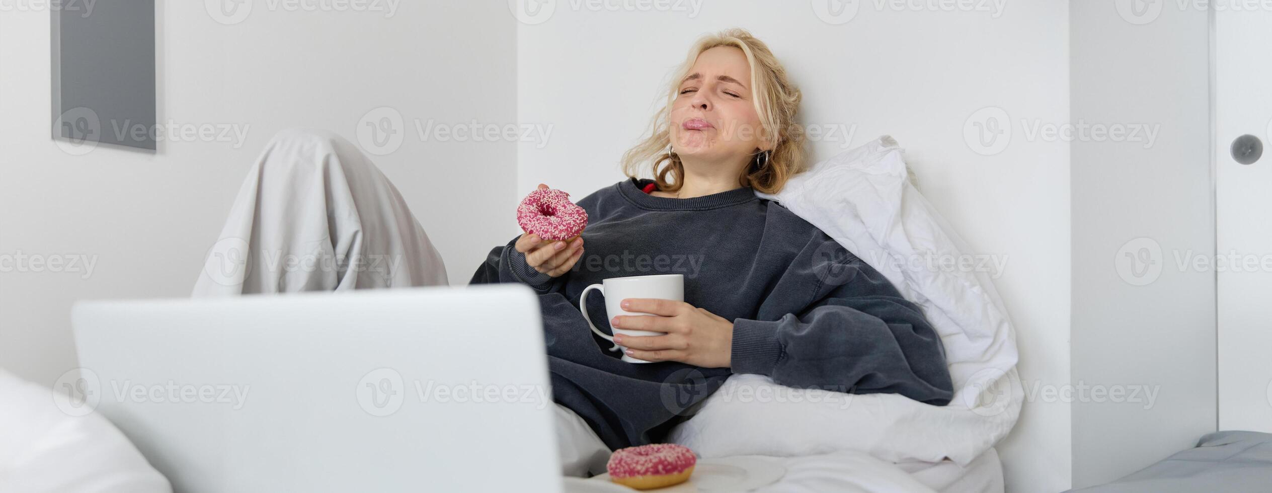 Portrait of sad and depressed woman, feeling heartbroken, lying in bed with comfort food, eating doughnut, crying from upsetting movie, watching something on laptop photo
