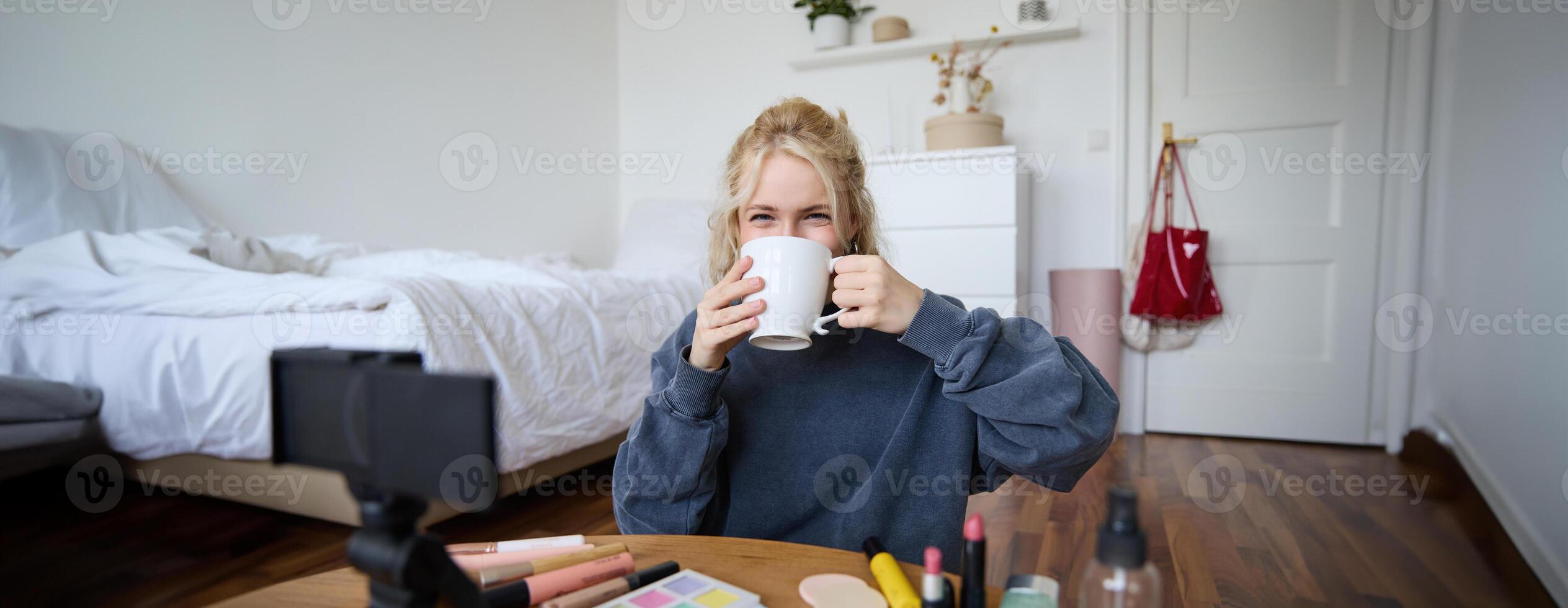 Image of young woman, makeup vlogger, sitting in bedroom with digital camera, drinking tea and talking, creating lifestyle , social media content photo
