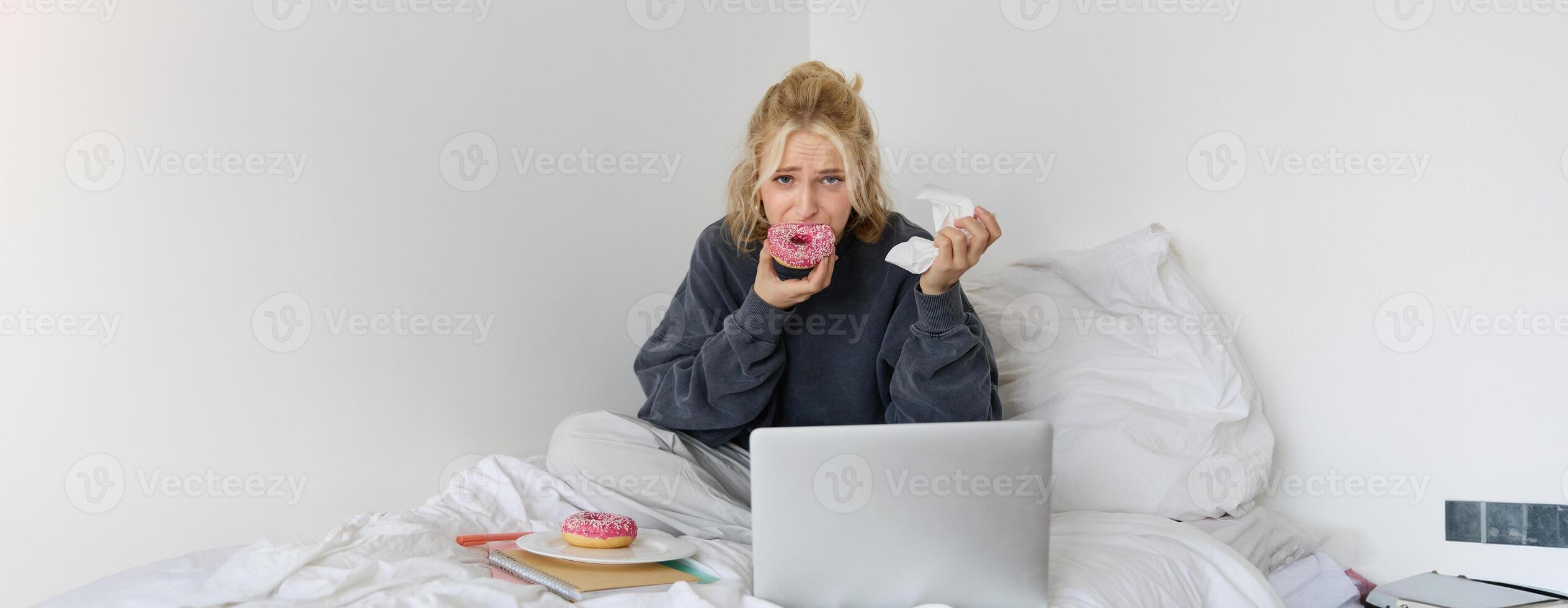 Portrait of sad woman crying, eating doughnut, wiping tears off, looking at something upsetting on laptop screen, sitting on a bed photo